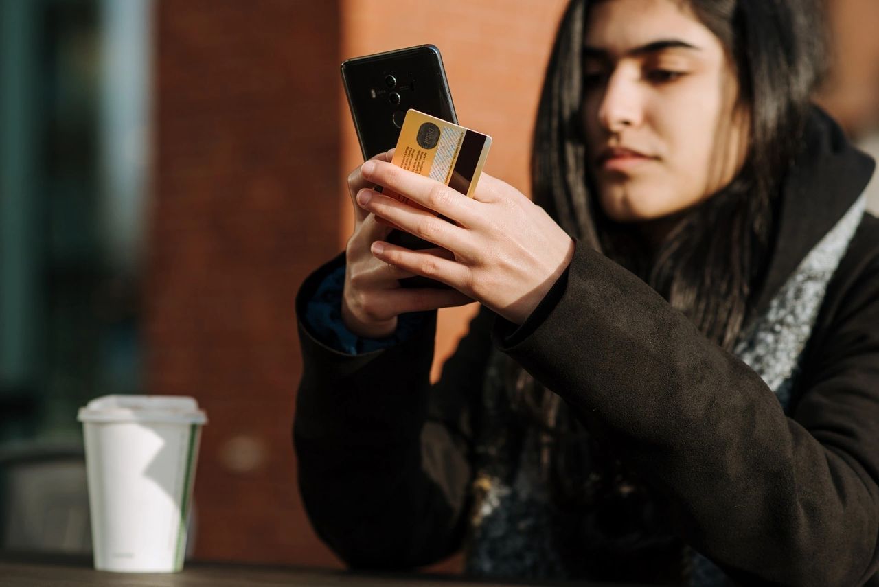 A woman on a coffee shop with her coffee on the side, holding her phone and credit card.