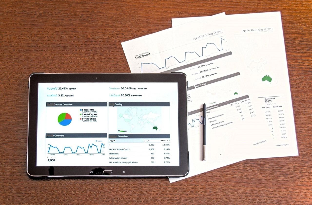 A photo of a tablet device with reports and pie chart, along with printed papers on the right, with graphs and financial report. 